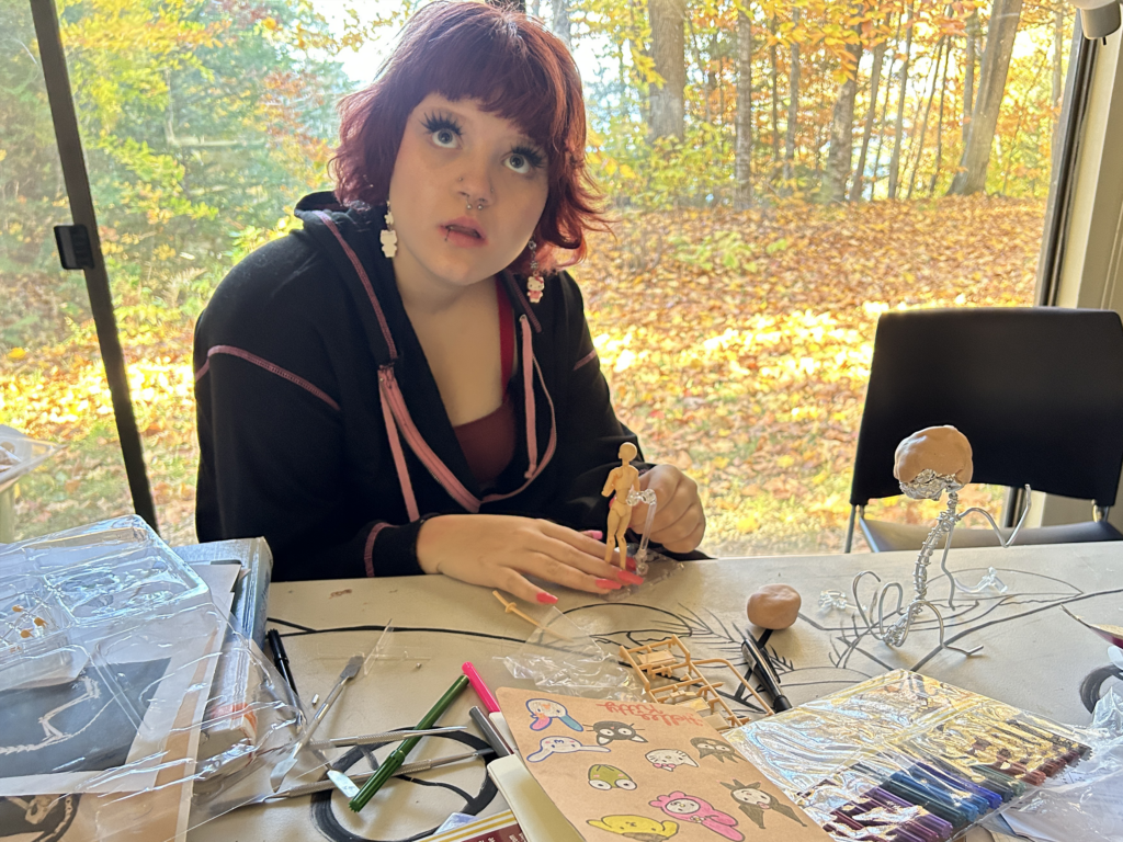 Young woman looking up at the photographer as she poses a small wooden figure. There is a sculpture in progress made of wire and clay on a table littered with art supplies. The background is a large window looking out on red and gold leaves in a wooded area on a sunny day.