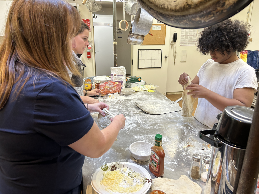 Young people making pizza dough in a kitchen with a teacher observing.